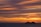 A large cruise ship travels on the ocean against a backdrop of a vibrant sunset. The sky is filled with streaks of orange, yellow, and purple clouds, creating a dramatic and serene atmosphere.