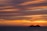 A large cruise ship travels on the ocean against a backdrop of a vibrant sunset. The sky is filled with streaks of orange, yellow, and purple clouds, creating a dramatic and serene atmosphere.
