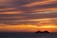 A large cruise ship travels on the ocean against a backdrop of a vibrant sunset. The sky is filled with streaks of orange, yellow, and purple clouds, creating a dramatic and serene atmosphere.