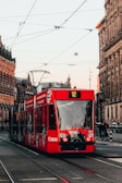 A red tram marked with advertising moves along a city street surrounded by historic buildings. Overhead wires and tram tracks are prominent. Pedestrians and urban infrastructure are visible, indicating a lively urban environment.