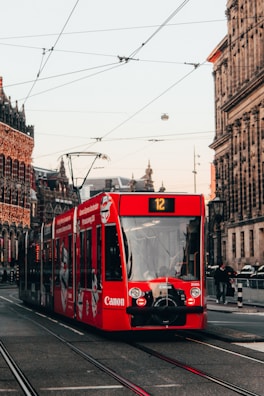 A red tram marked with advertising moves along a city street surrounded by historic buildings. Overhead wires and tram tracks are prominent. Pedestrians and urban infrastructure are visible, indicating a lively urban environment.