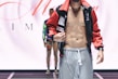A male model is wearing a fashion-forward outfit featuring grey pants and a colorful, open jacket revealing a toned torso. The background includes another person standing in a similar pose on a runway.