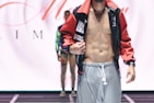 A male model is wearing a fashion-forward outfit featuring grey pants and a colorful, open jacket revealing a toned torso. The background includes another person standing in a similar pose on a runway.