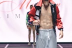 A male model is wearing a fashion-forward outfit featuring grey pants and a colorful, open jacket revealing a toned torso. The background includes another person standing in a similar pose on a runway.