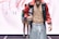 A male model is wearing a fashion-forward outfit featuring grey pants and a colorful, open jacket revealing a toned torso. The background includes another person standing in a similar pose on a runway.