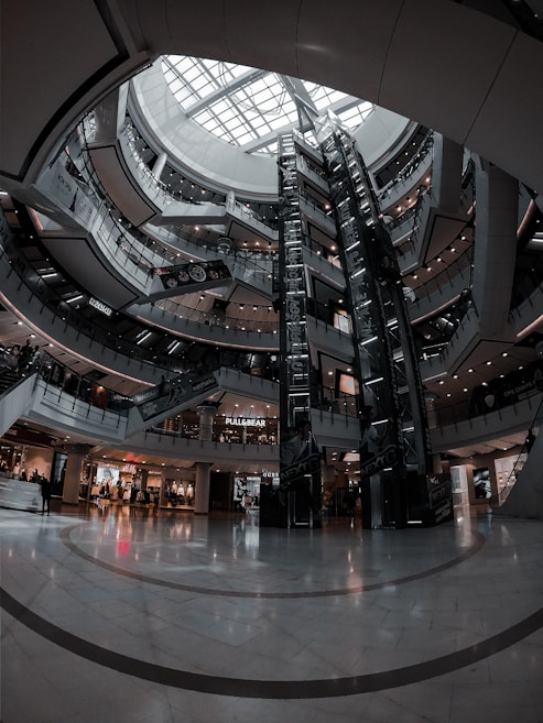 A large, multi-level shopping mall with a modern architectural design. The central area features a tall glass ceiling that allows natural light to illuminate the space. There are multiple floors visible with escalators and various storefronts that include popular fashion brands.