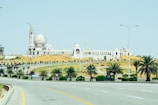 A large mosque with a prominent white dome and multiple minarets under construction. The building is surrounded by a desert landscape with patches of greenery, including several palm trees lining the road in the foreground. The road is wide and mostly empty, curving slightly with streetlights lining the sides under a clear blue sky.