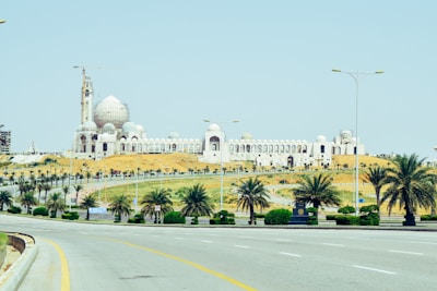 A large mosque with a prominent white dome and multiple minarets under construction. The building is surrounded by a desert landscape with patches of greenery, including several palm trees lining the road in the foreground. The road is wide and mostly empty, curving slightly with streetlights lining the sides under a clear blue sky.