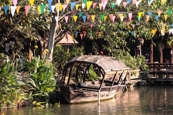 A rustic wooden boat with a thatched roof is moored along a small river, surrounded by lush greenery and vibrant flowers. Multicolored triangular flags are strung above, creating a festive atmosphere. Several wooden structures can be seen in the background, partially obscured by large trees.