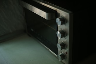 A close-up image of a stainless steel countertop oven with four control knobs is seen. The knobs are aligned vertically, and the oven door has a large handle. The surface appears clean and slightly reflective.