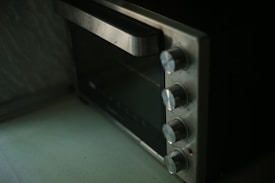 A close-up image of a stainless steel countertop oven with four control knobs is seen. The knobs are aligned vertically, and the oven door has a large handle. The surface appears clean and slightly reflective.
