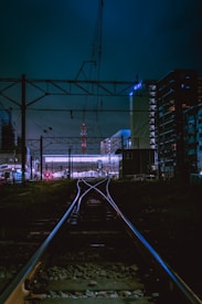 A railway track at night with the tracks leading into a station area that is illuminated with bright lights. Overhead electrical lines are visible, and there are buildings on both sides of the tracks. The atmosphere is urban with a dark and moody ambiance.
