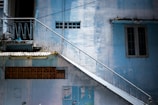 A worn-down exterior of a building with light blue walls, featuring a slanted metal staircase with railings. The building has small windows and a decorative brick element. The wall surfaces show signs of aging, with peeling paint and some grime.