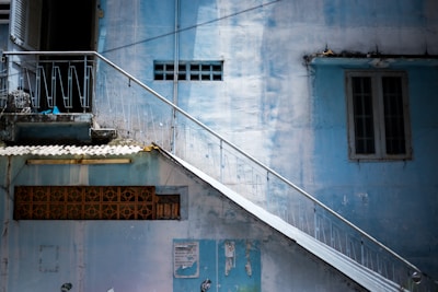 A worn-down exterior of a building with light blue walls, featuring a slanted metal staircase with railings. The building has small windows and a decorative brick element. The wall surfaces show signs of aging, with peeling paint and some grime.
