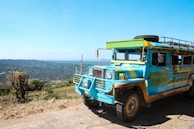 A brightly colored public transport vehicle with a distinctive style is parked on a paved area. The background shows a scenic landscape with rolling hills under a clear blue sky.