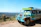 A brightly colored public transport vehicle with a distinctive style is parked on a paved area. The background shows a scenic landscape with rolling hills under a clear blue sky.