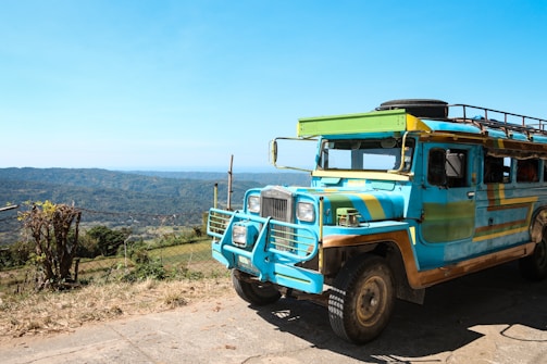 A brightly colored public transport vehicle with a distinctive style is parked on a paved area. The background shows a scenic landscape with rolling hills under a clear blue sky.