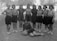 A group of nine women wearing early 20th-century athletic clothing, consisting of shorts, long-sleeved shirts, and patterned socks, stand together on a dirt field. One woman is sitting on the ground in front, resting her arm on a soccer ball. The background includes trees and indistinct buildings, suggesting an outdoor setting.