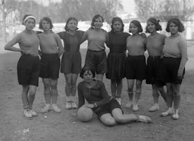 A group of nine women wearing early 20th-century athletic clothing, consisting of shorts, long-sleeved shirts, and patterned socks, stand together on a dirt field. One woman is sitting on the ground in front, resting her arm on a soccer ball. The background includes trees and indistinct buildings, suggesting an outdoor setting.