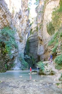 A serene scene features a narrow gorge with high rocky walls covered in patches of greenery. A small waterfall can be seen at the center, cascading down into a shallow, clear pool of water at the bottom. A few people are present, exploring and enjoying the natural surroundings. They appear to be hiking along the rocky terrain.