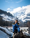 woman in blue jacket and blue backpack sitting on rock near snow covered mountain during daytime