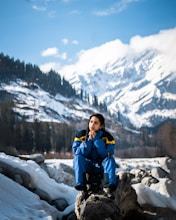 woman in blue jacket and blue backpack sitting on rock near snow covered mountain during daytime