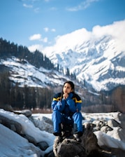 woman in blue jacket and blue backpack sitting on rock near snow covered mountain during daytime