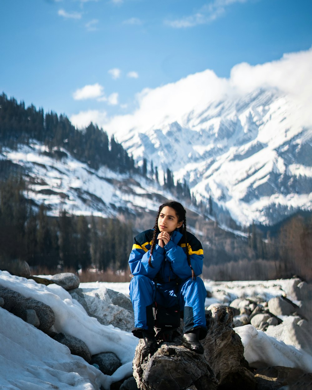 woman in blue jacket and blue backpack sitting on rock near snow covered mountain during daytime