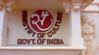 A decorative wall features a circular emblem with a stylized Om symbol in white on a red background accompanied by the text 'Ministry of Culture Govt. of India'. To the right, there is a detailed sculpture of a traditional Indian figure carved into the sandstone.