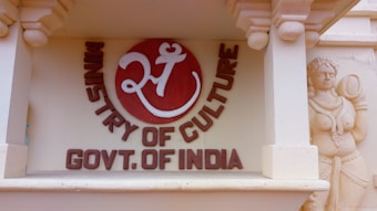 A decorative wall features a circular emblem with a stylized Om symbol in white on a red background accompanied by the text 'Ministry of Culture Govt. of India'. To the right, there is a detailed sculpture of a traditional Indian figure carved into the sandstone.