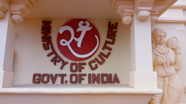 A decorative wall features a circular emblem with a stylized Om symbol in white on a red background accompanied by the text 'Ministry of Culture Govt. of India'. To the right, there is a detailed sculpture of a traditional Indian figure carved into the sandstone.