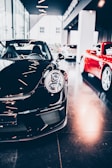 A sleek black sports car is prominently displayed indoors, surrounded by other luxury vehicles. The environment suggests a high-end showroom or dealership with bright lighting and modern architecture. The flooring is reflective, adding to the luxurious tone.