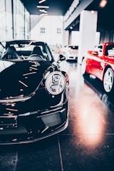 A sleek black sports car is prominently displayed indoors, surrounded by other luxury vehicles. The environment suggests a high-end showroom or dealership with bright lighting and modern architecture. The flooring is reflective, adding to the luxurious tone.