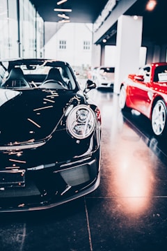 A sleek black sports car is prominently displayed indoors, surrounded by other luxury vehicles. The environment suggests a high-end showroom or dealership with bright lighting and modern architecture. The flooring is reflective, adding to the luxurious tone.