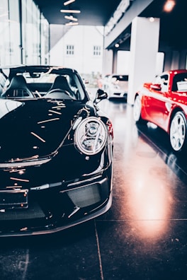 A sleek black sports car is prominently displayed indoors, surrounded by other luxury vehicles. The environment suggests a high-end showroom or dealership with bright lighting and modern architecture. The flooring is reflective, adding to the luxurious tone.