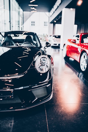 A sleek black sports car is prominently displayed indoors, surrounded by other luxury vehicles. The environment suggests a high-end showroom or dealership with bright lighting and modern architecture. The flooring is reflective, adding to the luxurious tone.