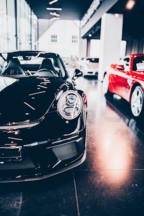 A sleek black sports car is prominently displayed indoors, surrounded by other luxury vehicles. The environment suggests a high-end showroom or dealership with bright lighting and modern architecture. The flooring is reflective, adding to the luxurious tone.