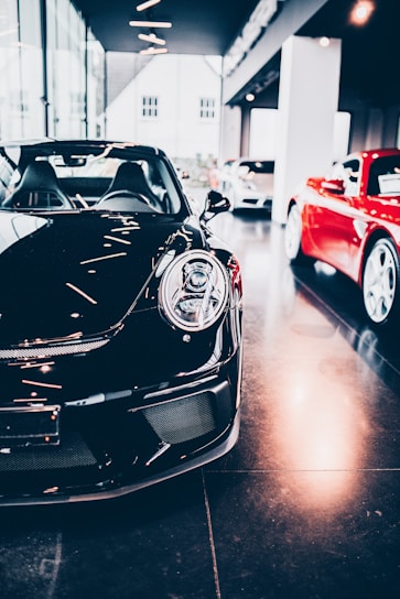 A sleek black sports car is prominently displayed indoors, surrounded by other luxury vehicles. The environment suggests a high-end showroom or dealership with bright lighting and modern architecture. The flooring is reflective, adding to the luxurious tone.
