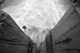 Black and white photograph capturing a dynamic view of ocean waves crashing between two wooden structures. The water appears to be in motion, possibly due to long exposure, creating a flowing and blurred effect. The wooden beams are weathered, suggesting they are part of a jetty or pier. Two metal bolts protrude from the wooden planks, indicating some form of construction or reinforcement.