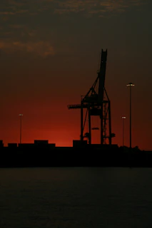 A towering ship-to-shore crane silhouetted against a sunset over a bustling container terminal.