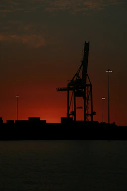 A towering ship-to-shore crane silhouetted against a sunset over a bustling container terminal.
