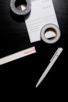 A black surface holds a white pen, two rolls of decorative tape, and a notepad labeled 'IDEA DIARY' with sections for 'Start', 'Volume', and more. The tape has a blue and white pattern, while the notepad is partly underneath one roll of tape.