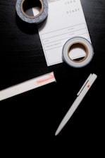 A black surface holds a white pen, two rolls of decorative tape, and a notepad labeled 'IDEA DIARY' with sections for 'Start', 'Volume', and more. The tape has a blue and white pattern, while the notepad is partly underneath one roll of tape.