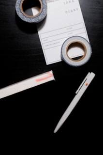 A black surface holds a white pen, two rolls of decorative tape, and a notepad labeled 'IDEA DIARY' with sections for 'Start', 'Volume', and more. The tape has a blue and white pattern, while the notepad is partly underneath one roll of tape.