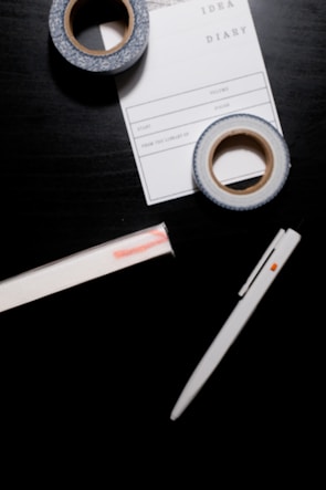 A black surface holds a white pen, two rolls of decorative tape, and a notepad labeled 'IDEA DIARY' with sections for 'Start', 'Volume', and more. The tape has a blue and white pattern, while the notepad is partly underneath one roll of tape.