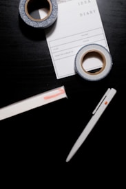 A black surface holds a white pen, two rolls of decorative tape, and a notepad labeled 'IDEA DIARY' with sections for 'Start', 'Volume', and more. The tape has a blue and white pattern, while the notepad is partly underneath one roll of tape.