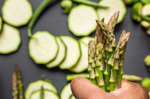 A hand holds a bunch of fresh asparagus spears over a dark surface. In the background, there are slices of zucchini and a whole green chili scattered around.