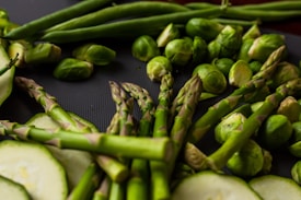 A close-up of fresh green vegetables, including asparagus spears, Brussels sprouts, zucchini slices, and green beans, arranged on a dark surface. The vibrant green hues dominate the composition, showcasing a variety of textures and shapes.