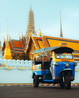 A colorful three-wheeled tuk-tuk parked in front of a grand temple with ornate golden spires and intricate architectural details.