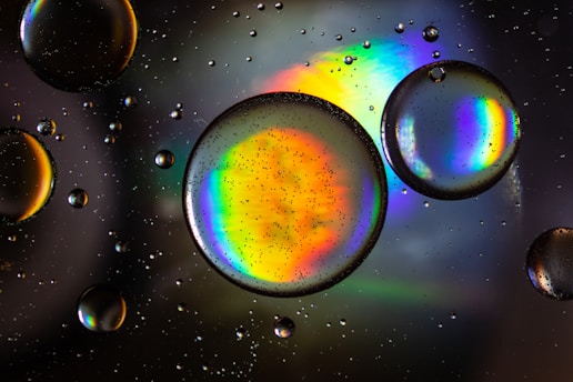 Large transparent bubbles are floating in a liquid, with a colorful rainbow spectrum reflected inside them. Smaller bubbles are dispersed throughout the background, creating a mesmerizing visual effect.